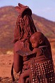 Comme beaucoup de femmes Africaines, l'enfant est maintenu sur le dos à l'aide de ce "porte bébé", harnais  en peau de chèvre,  dont les sangles sont nouées sur la poitrine.
Cette femme porte la coiffe qu'elle a mise pour son mariage et qui lui vient de sa mère. Portage d'un enfant sur le dos. Harnais en peau de chèvre. KAOKOLAND.NAMIBIE 